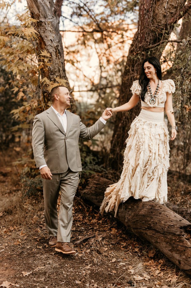 A couple in vintage western attire poses playfully on a fallen tree trunk.