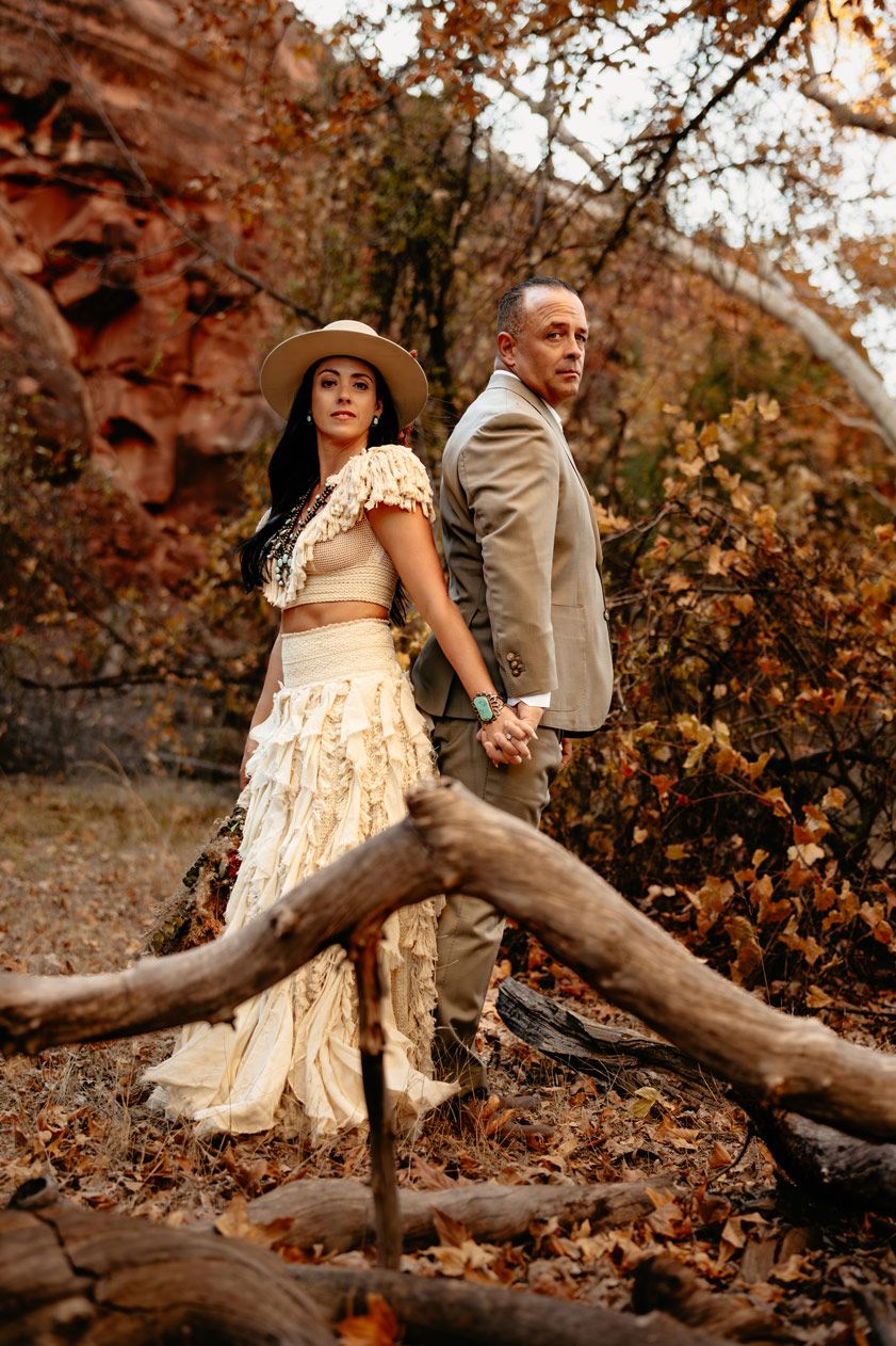 A stylish couple in western wedding attire poses against a fallen tree in autumn woods.