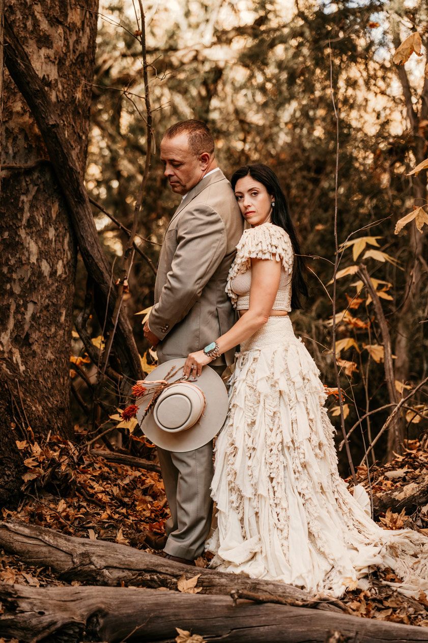 A couple in vintage western attire stands back-to-back in a wooded setting.