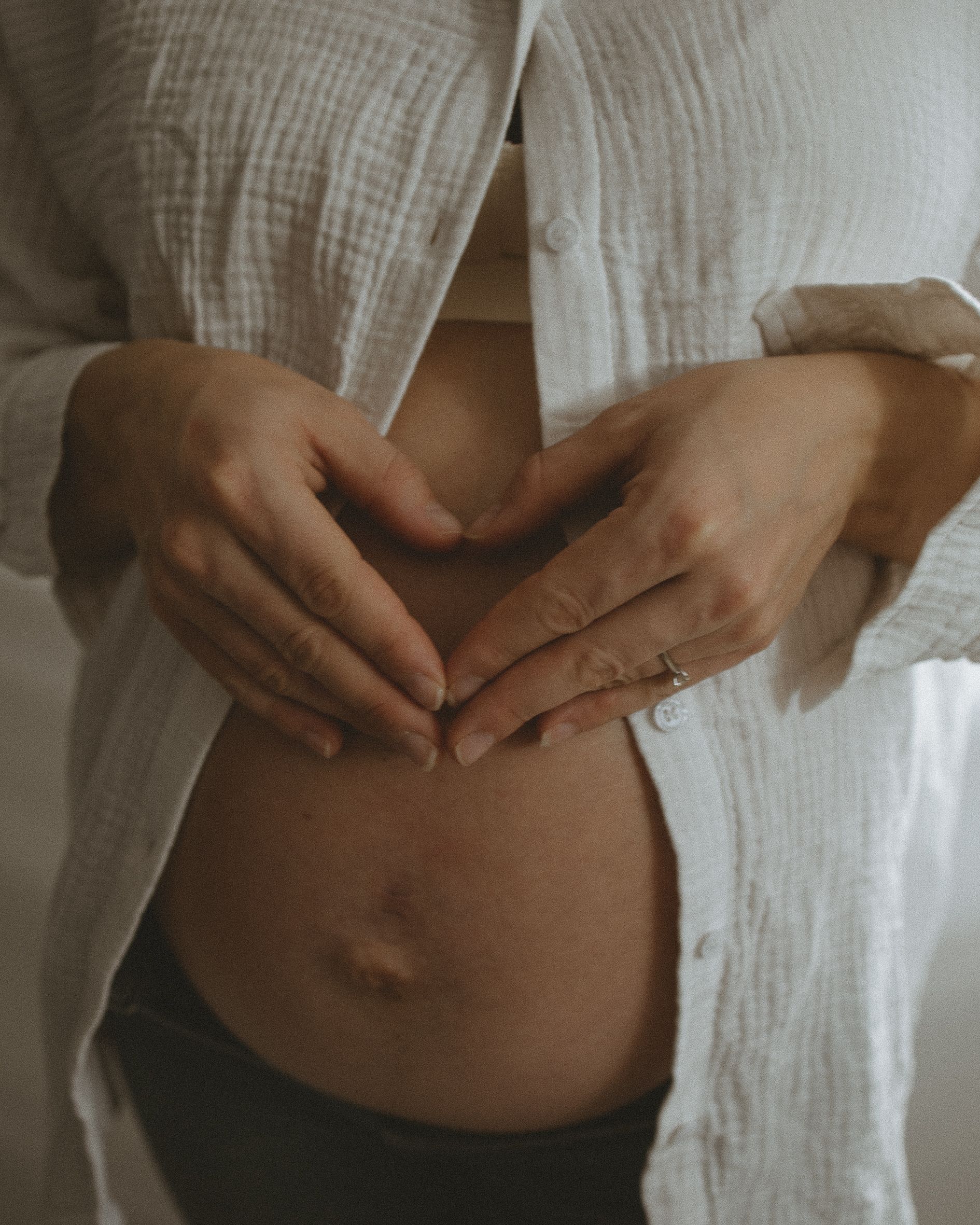 Hands form a heart shape over a pregnant belly in soft natural light.