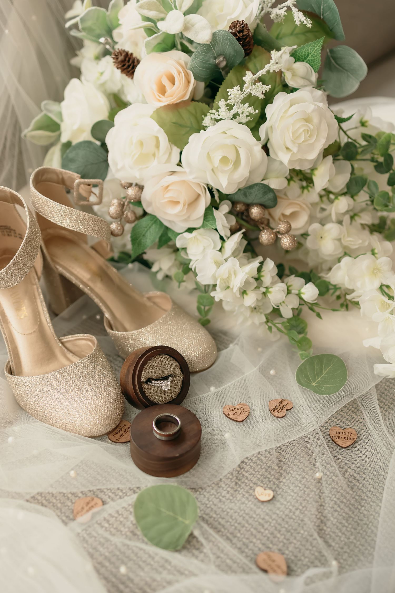 Wedding details with glittery gold heels, white rose bouquet with eucalyptus, and wooden ring boxes on delicate tulle fabric.