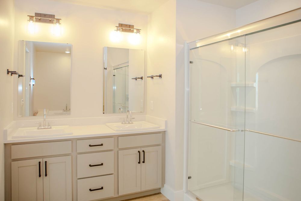 Modern white bathroom with dual sink vanity, bronze hardware, and glass shower enclosure with white tile surround.