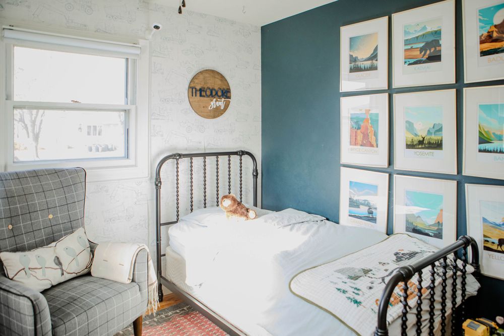 A cozy bedroom with a black metal bed frame, gray armchair, and colorful coastal artwork displayed on a teal accent wall.