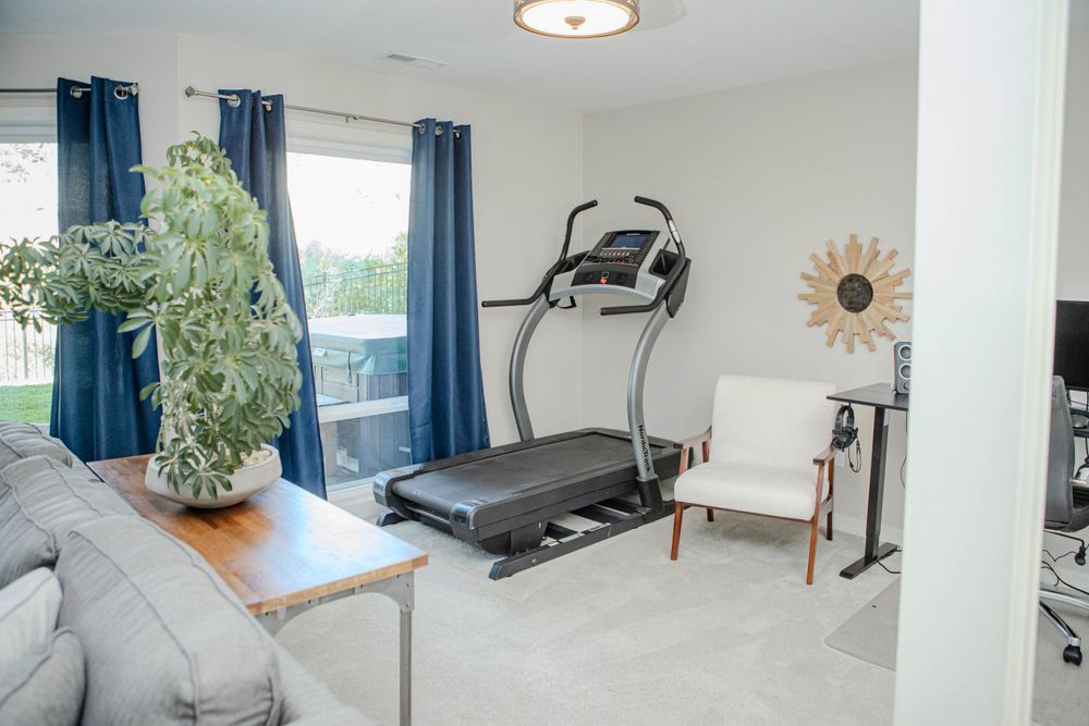 Modern home gym setup in bright room with treadmill, blue curtains, gray sofa, and potted plant by window.