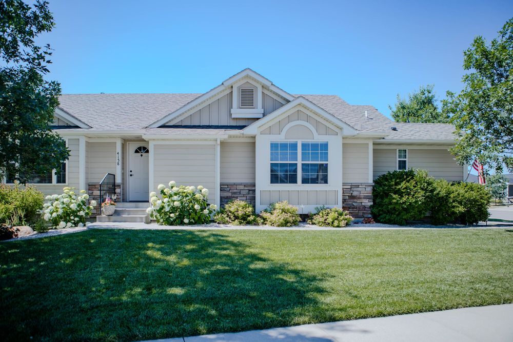 Single story ranch home with white siding, black shutters and colorful flower gardens on a manicured green lawn.