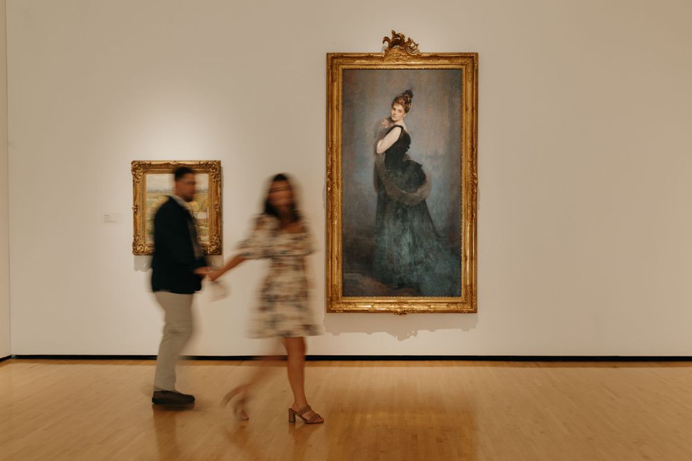 Motion-blurred figures view classical paintings in an art gallery with wooden floors.