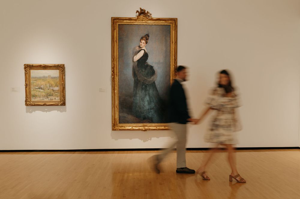 Blurred visitors walk past ornate framed paintings in a museum gallery.