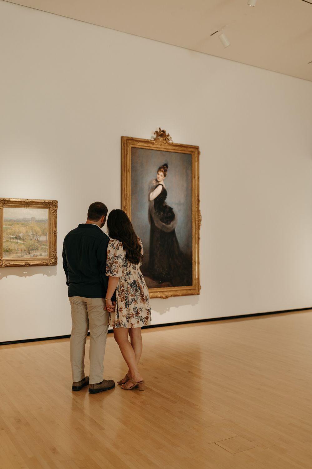 A couple observes a large portrait painting of a woman in a Victorian dress at an art museum.