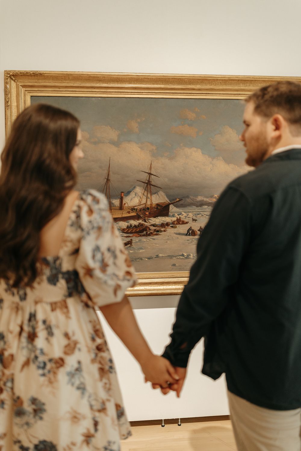 Couple holds hands while viewing a maritime painting in an art gallery.