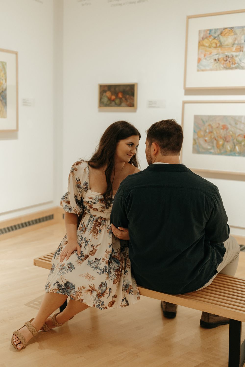 A couple shares a quiet moment on a wooden bench in an art gallery.