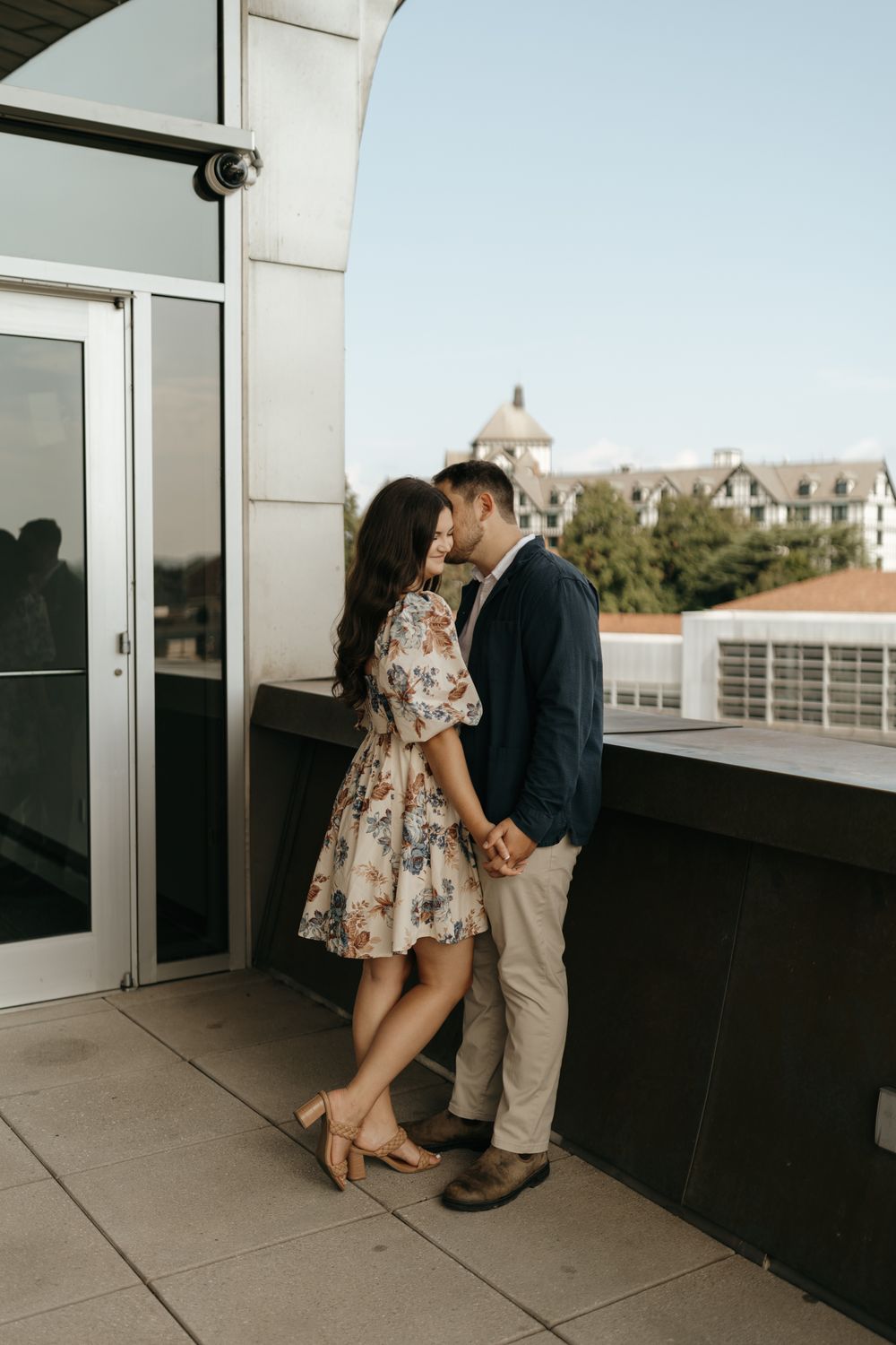 A romantic couple embraces on a balcony with a floral dress and navy blazer against an architectural backdrop.