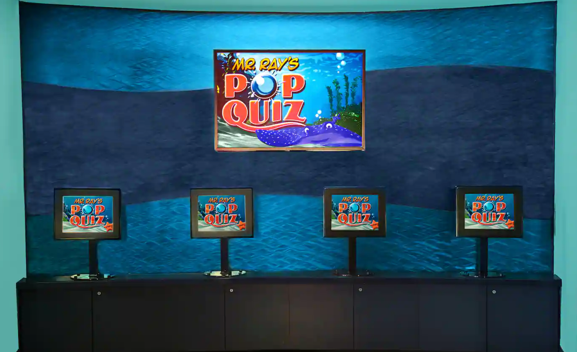 A gaming arcade with multiple TV screens mounted on a blue-lit wall displaying video game content.