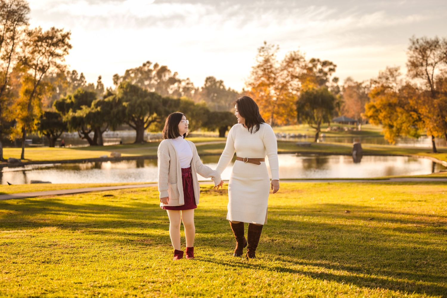 Two people holding hands while walking together on a grassy hill by a lake during golden sunset hour.