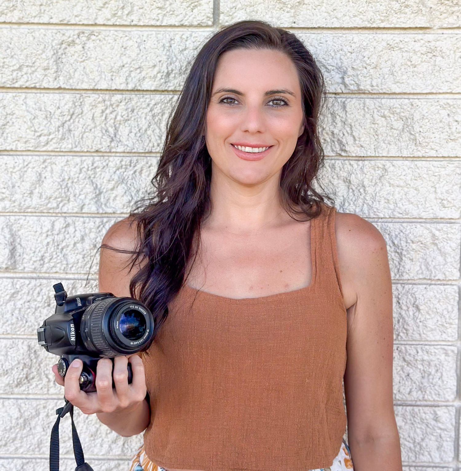 Professional photographer in brown tank top holding black DSLR camera against white brick wall.