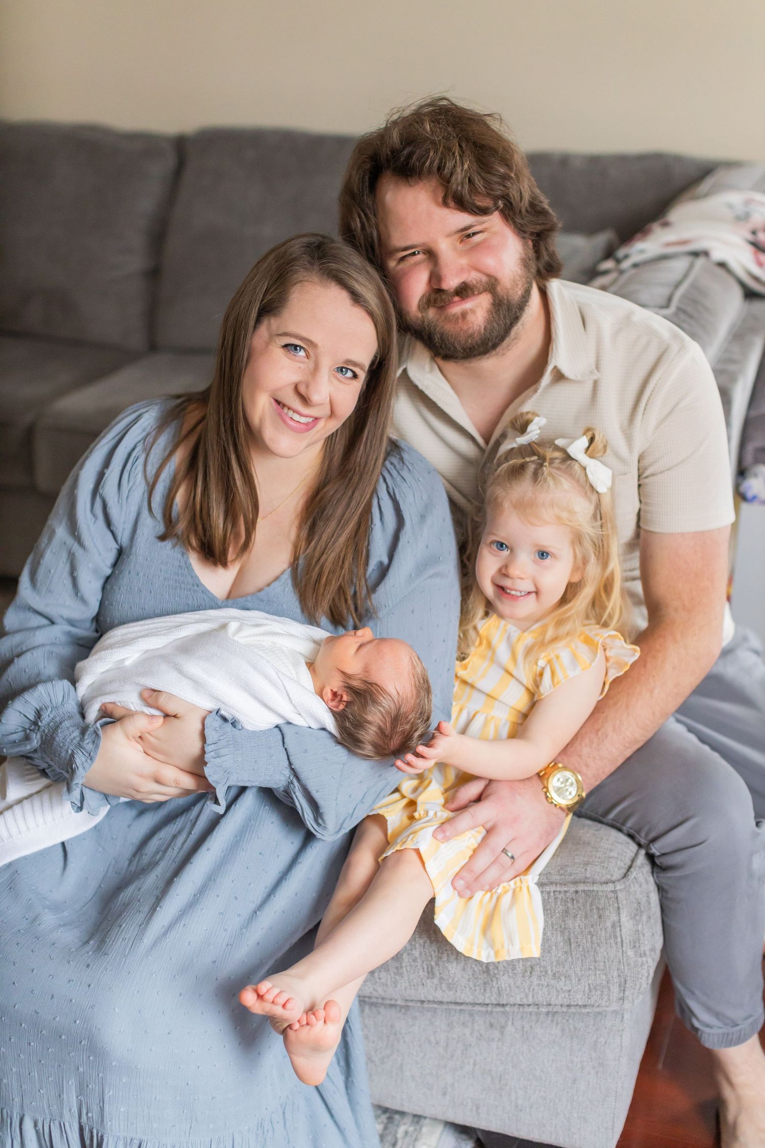 A cheerful family relaxes together on a grey couch, with a newborn baby and a young child in a yellow dress.