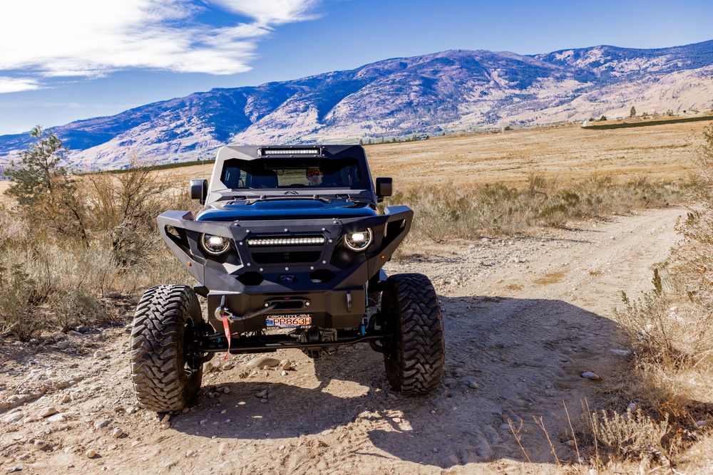 A dark blue Jeep Gladiator drives along a rugged mountain trail with snow-capped peaks in the background.