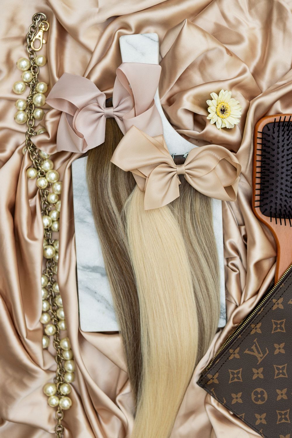 Blonde hair extensions displayed on silk champagne fabric with Louis Vuitton accessories and styling tools.