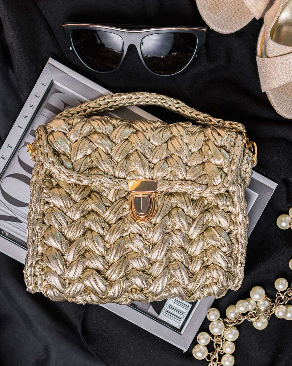 Stylish woven straw handbag displayed with sunglasses, pearls and fashion magazine on black background.