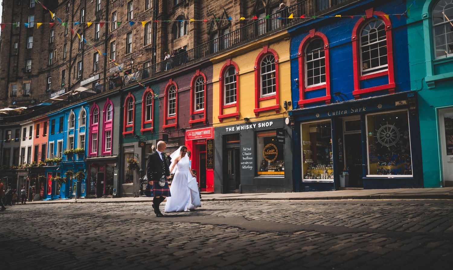 A figure in white walks past colorful storefronts with arched windows on a historic cobblestone street in Edinburgh. City Chambers Wedding