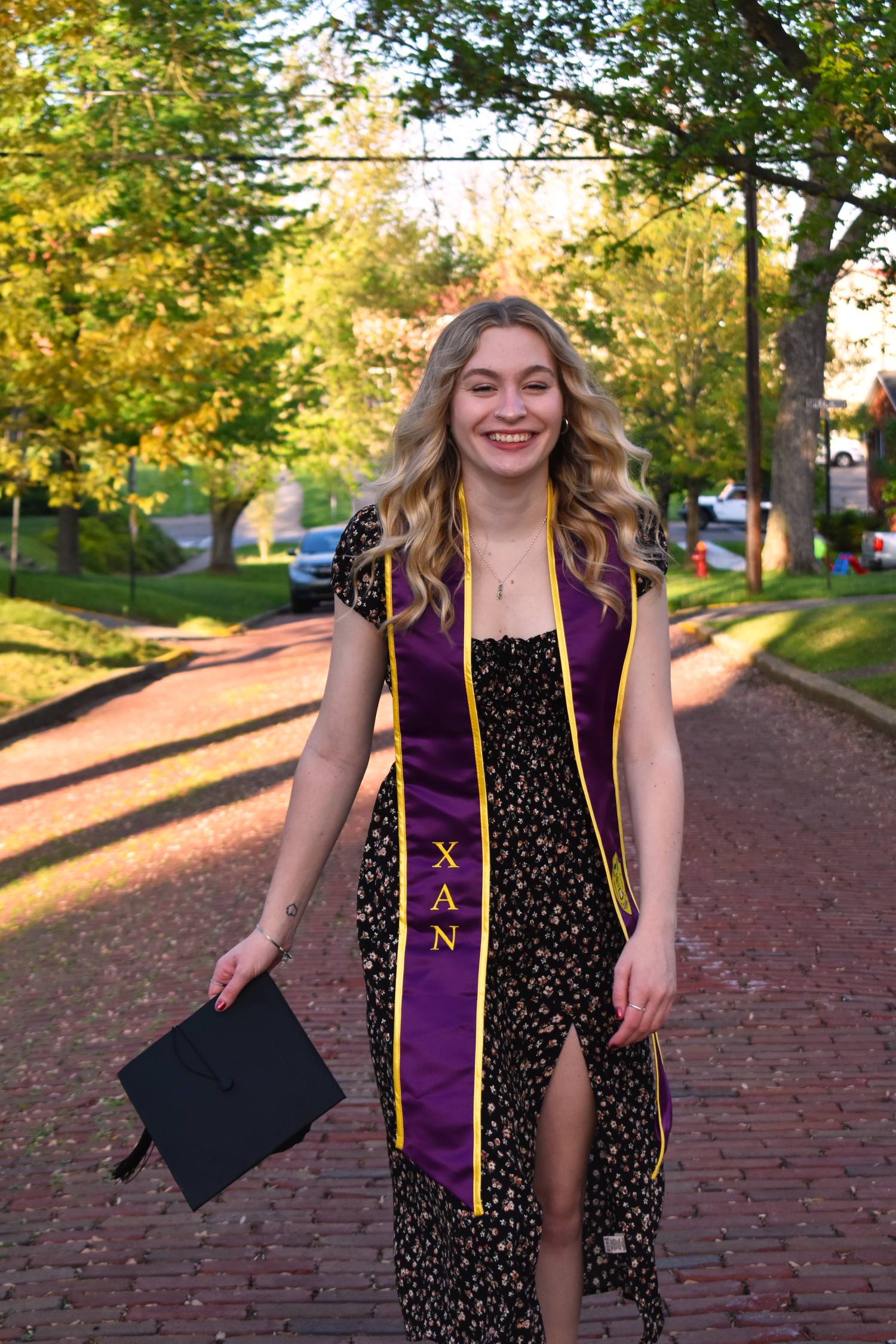Graduate wearing a purple Chi Nu stole and floral dress holds a cap while standing on a brick path during golden hour.