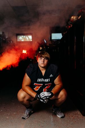 High school football player number 77 in Indians jersey crouches in a locker room with dramatic red smoke behind him.