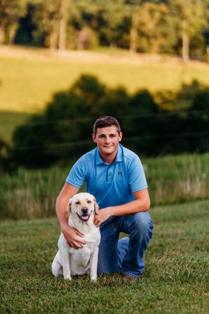 Smiling young man in blue polo kneeling beside a yellow Labrador Retriever on green grass.