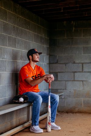 Baseball player in orange RRHS jersey sitting in dugout leaning on bat with glove and ball.