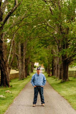 Man standing on tree-lined gravel farm driveway with hands in pockets on a cloudy day.