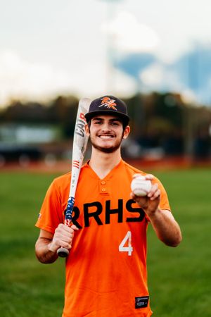 Smiling RRHS number 4 baseball player holding bat and ball in orange uniform outdoors.