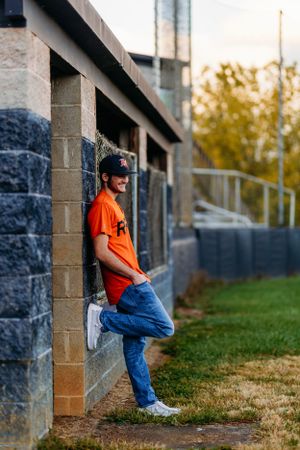 Teen in orange RRHS baseball jersey leaning against concrete wall at a baseball stadium.