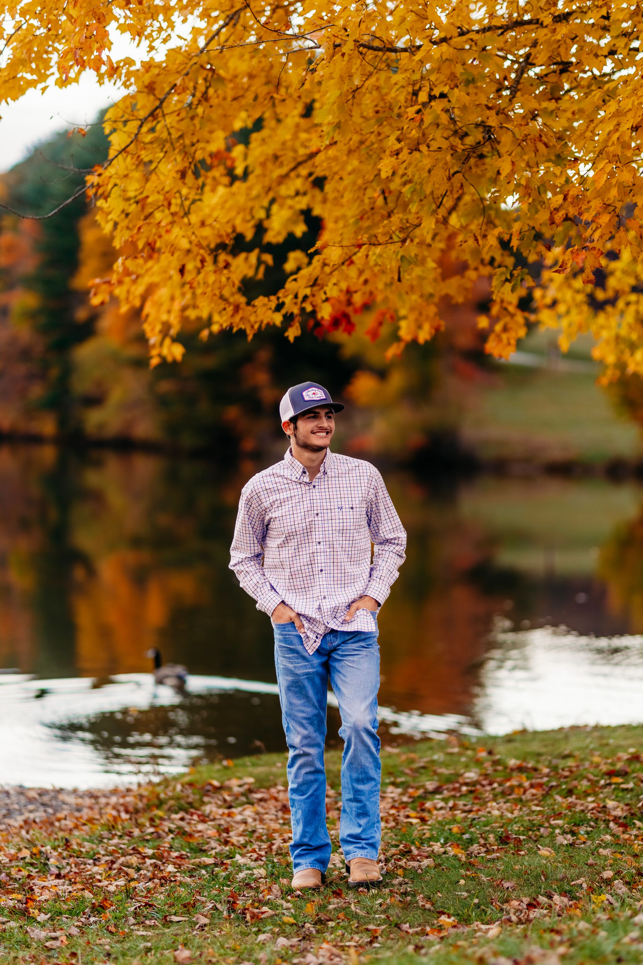 Young man in plaid shirt and jeans standing near a pond under vibrant autumn foliage.