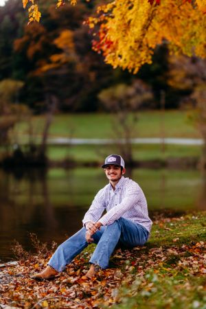 Smiling young man sitting by a pond surrounded by colorful fall foliage and leaves.