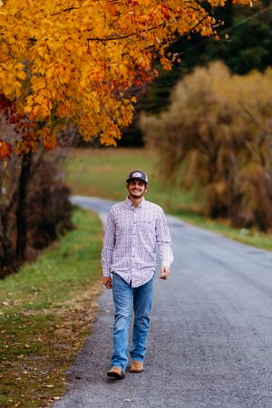 Smiling young man in plaid shirt walking down a road under vibrant autumn foliage.