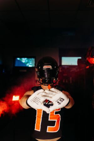 Football player wearing number 15 jersey and helmet holds a ball in a dark dramatic locker room with red smoke.