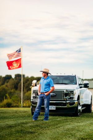 Man in blue polo and cowboy hat standing beside white Ford truck with American and Marine Corps flags.