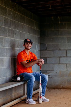 Smiling baseball player in orange jersey sitting in dugout holding a bat with glove nearby.