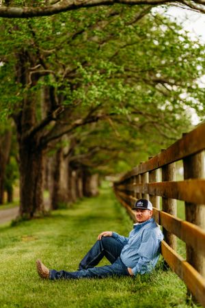 Young man in blue shirt and cap relaxing against wooden fence on lush green farm lane.