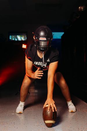 High school football player in Indians helmet crouches over a football in a dark dramatic tunnel setting.