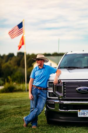 Man in cowboy hat and blue polo leaning on Ford truck grille with American flag in background.