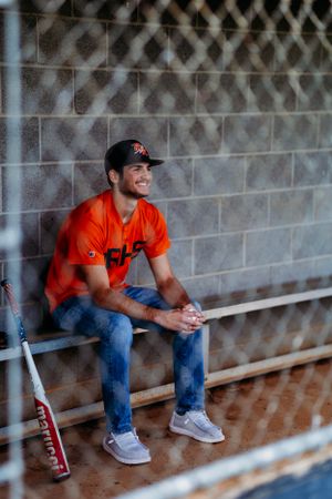 Baseball player in orange jersey smiling in dugout with Marucci bat leaning against wall.
