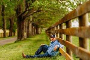 Man in blue plaid shirt sitting against rustic wooden fence along tree-lined farm path.