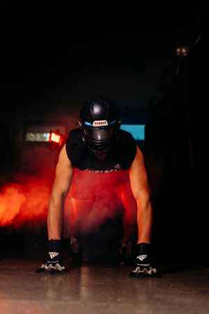 American football player in black helmet crouches in a dramatic pose surrounded by red smoke in a dark tunnel.