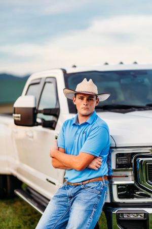 Young man in blue polo, cowboy hat, and jeans leaning against white Ford pickup truck outdoors.