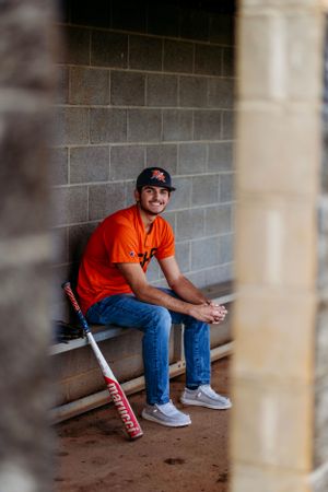 Smiling baseball player in orange jersey sitting in dugout with Marucci bat leaning nearby.