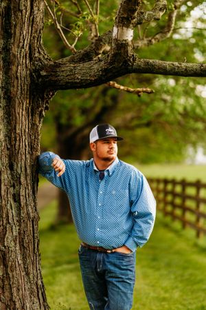 Young man in blue patterned shirt and cap leaning against a large tree trunk by wooden fence.
