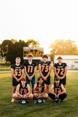 Seven high school Indians football players pose together on a field at golden hour with a scoreboard behind them.