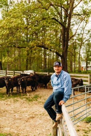 Farmer sitting on fence rail with black Angus cattle in background on a rural farm.