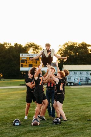 Football players lifting a teammate in the air on a green field at sunset with a scoreboard in the background.