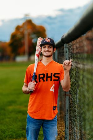 Baseball player in orange RRHS #4 jersey holding a Marucci bat against a chain-link fence.