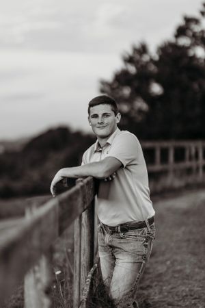 Black and white portrait of teenage boy leaning on wooden fence in rural countryside.